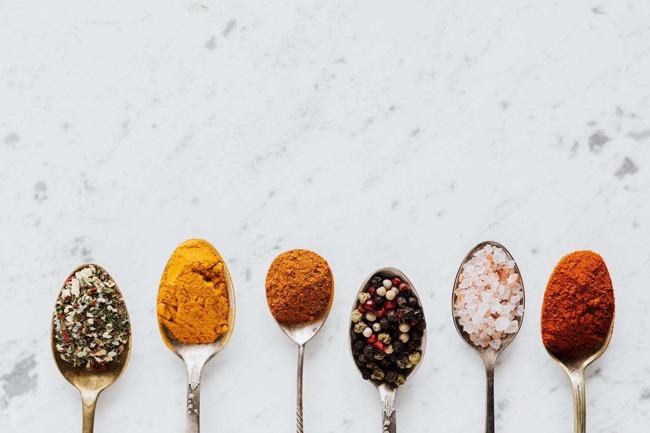 Top view of assorted aromatic condiments arranged together in metal spoons on marble surface used for delicious gourmet meal cooking