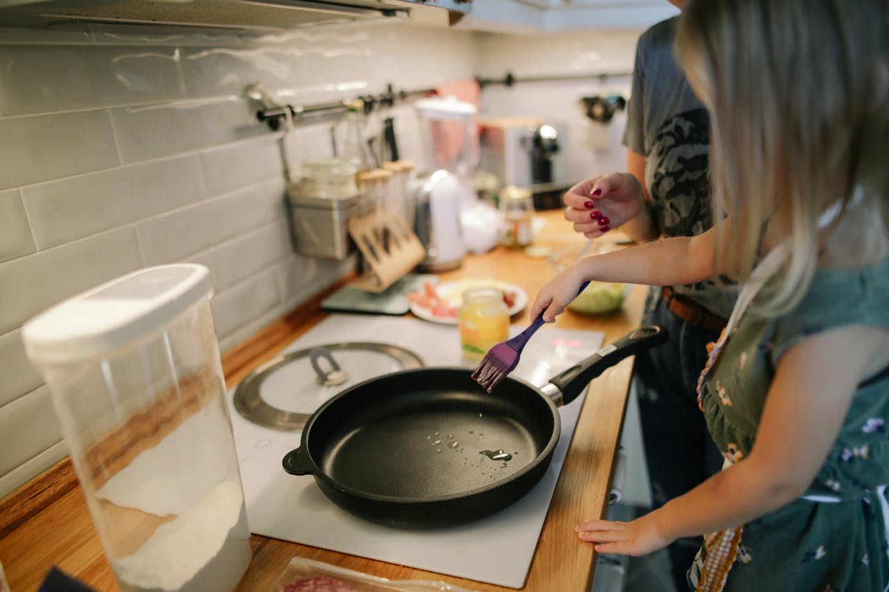 Young girl cooking with a frying pan under adult supervision in a modern kitchen.